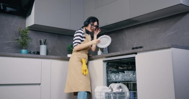 Νεαρή ελκυστική γυναίκα με ποδιά και ακουστικά πλένει πιάτα και χορεύει, αισθάνεται ευτυχισμένη στην κουζίνα — Αρχείο Βίντεο