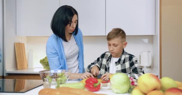 Encantadora mãe de cabelos escuros que ajuda seu filho de 12 anos a cortar pimenta para salada de legumes saborosa enquanto prepara o jantar em família junto com seu marido na cozinha moderna — Vídeo de Stock