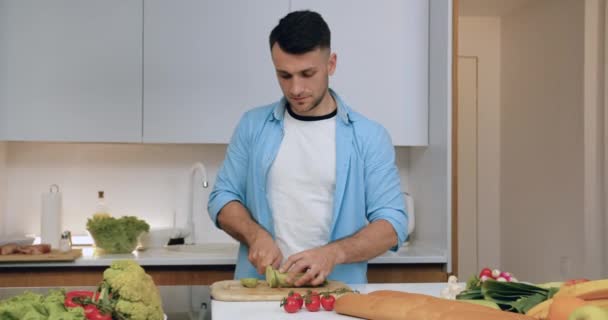 Vooraanzicht van knappe tevreden bekwame man die zucchini snijden op snijplank met mes en dan te kijken naar de camera met oprechte glimlach — Stockvideo
