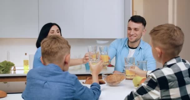 Портрет любящей современной счастливой семьи с детьми, которые сидят за обеденным столом в уютной кухне и звон стаканов с апельсиновым соком во время завтрака утром — стоковое видео
