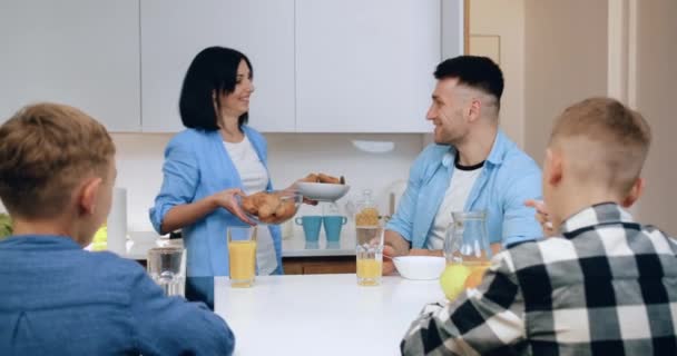 Cukup tersenyum bahagia ibu berambut hitam menempatkan di atas croissant meja dan oatcookie di belakang yang duduk dua putra dan suaminya, konsep sarapan keluarga — Stok Video