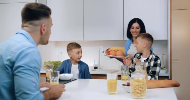 Atractivo padre y dos niños sentados en la mesa y comiendo cereales cuando sonríe madre morena lleva un plato grande con frutas y poner en la mesa — Vídeos de Stock