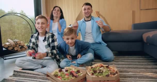 Idilio familiar donde los padres emotivos sonrientes y guapos celebran la victoria de los niños en el videojuego en la sala de estar y dos sabrosas pizzas que comerán más tarde — Vídeo de stock