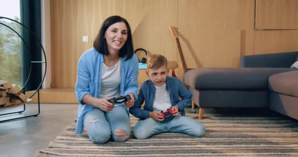 Fermecător fericit zâmbind mama cu părul negru stând pe podea împreună cu fiul ei și jucând jocuri video folosind joystick-uri atunci când petrec timpul liber comun acasă — Videoclip de stoc