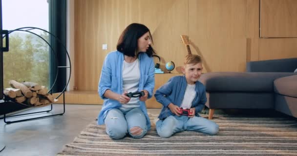 Framifrån av attraktiva glada leende mor och hennes lilla son som sitter på golvet och njuter av videospel med hjälp av gamepads hemma — Stockvideo