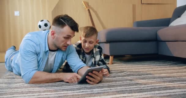 英俊而积极的现代父亲和他的儿子一起躺在地板上，向他解释如何使用平板电脑，家庭休闲概念 — 图库视频影像