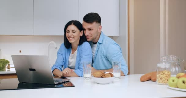 Привлекательная счастливая улыбающаяся супружеская пара машет руками, чтобы поздороваться со своим другом или родственником через компьютерный видеочат во время завтрака на кухне — стоковое видео