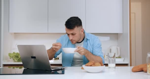 Atractivo satisfecho hombre moderno de 35 años sentado en la mesa de la cocina, comer cereales para el desayuno y ver videos divertidos en el ordenador portátil, de cerca — Vídeos de Stock