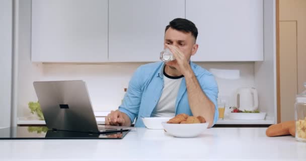 Çağdaş mutfakta kahvaltı yaparken süt içen ve dizüstü bilgisayarla çalışmaya devam eden hoş, konsantre genç bir adam. — Stok video