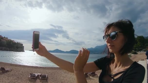 Vista lateral. Bonita joven en gafas de sol toma selfie en su teléfono inteligente en la isla Sveti Stefan en Montenegro — Vídeos de Stock