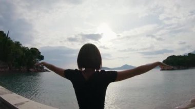 Genç bir kadın parkta yürüyor ve gün batımında inanılmaz deniz manzarasının önünde kollarını açmış denize yaklaşıyor..