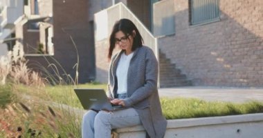 Laptop bilgisayar kullanan, notlar alan, kampüs dışında oturarak uzaktan eğitim alan çekici beyaz kadın öğrenci. Dışarıda.