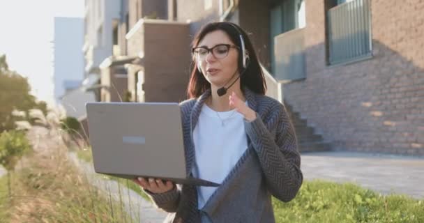 Mulher bastante ocupada de 30 anos com fones de ouvido e dispositivo de computador durante o trabalho on-line ou aprendizagem remota no laptop ao ar livre — Vídeo de Stock