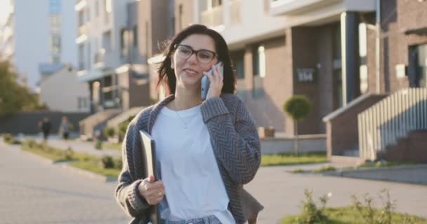 Pewna siebie i uśmiechnięta młoda kaukaska kobieta w okularach trzymająca laptopa podczas spaceru rozmawiając z najlepszym przyjacielem przez telefon komórkowy przy wietrznej słonecznej pogodzie — Wideo stockowe