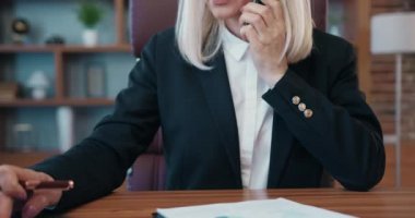 Beautiful smiling older businesswoman is talking on mobile phone and typing on laptop in modern office communicating with business partners. Discuss business issues on phone.