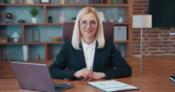 Portrait of beautiful smiling woman specialist in eyeglasses sitting at a desk in the office and looking at the camera — стоковое видео