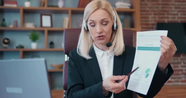 Elegant middle aged businesswoman sitting at office desk, showing financial report having conference call with client on laptop in office — ストック動画