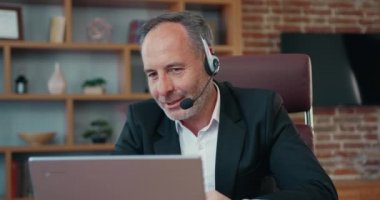 Mature businessman CEO holding video conference call with clients, wearing wireless headphones with microphone. Handsome worker having video call on laptop computer.