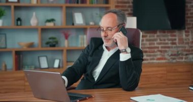 Bearded businessman CEO answering on phone call while using computer. Man office worker manager consulting client by telephone checking information on laptop screen.