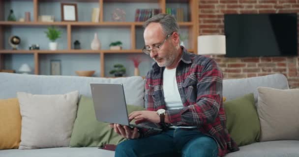 Handsome mature man working on laptop while sitting on a sofa in living room. Male learning using computer, making notes. Freelancer Working From Home. — Stok video