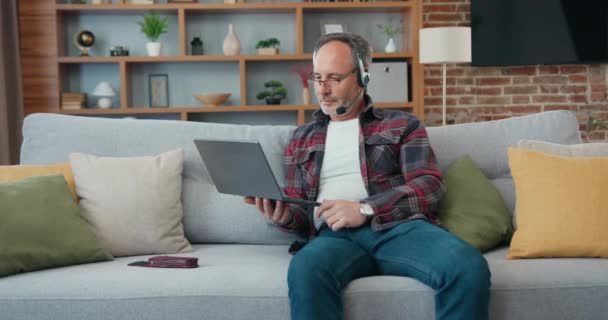 Middle aged man in headphones, talking video call, sitting on the sofa at home working. Mature male lying on the coach at the laptop makes an internet video conference call. — Αρχείο Βίντεο