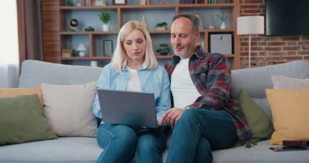 Feliz pareja de mediana edad mirando la pantalla del ordenador portátil sentado en el sofá en casa. Mujer y hombre en la sala de estar de búsqueda de Internet escribiendo en línea utilizando el ordenador viendo vídeo navegar por Internet. — Vídeos de Stock