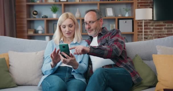Mature couple at home using smart phone together and smiling. Man and woman relaxing on sofa and shopping on internet, watching funny videos, use social media. — Video