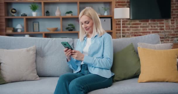 Smiling middle aged lady customer texting mobile sms message playing mobile games using social media apps at home. Wanita Millennial menghabiskan waktu di rumah dengan teknologi gadget sel. — Stok Video