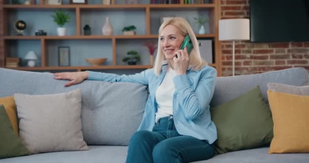Happy middle aged woman spend leisure alone at home relaxing on sofa, talking on smartphone. Blond famele at home make telephone call talk enjoy conversation. — Stok video