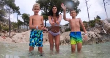Adriyatik Denizi 'nde tatilin tadını çıkaran iki oğluyla neşeli bir kadın. Sahilde neşeli bir aile baş parmaklarını sallıyor ve el sallıyor.
