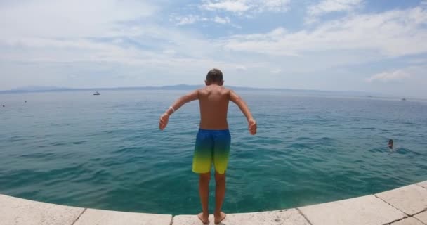 Молодий хлопчик у плавальних шортах занурюється з пірсу в прозоре блакитне море, каміння можна побачити на дні моря. Літні канікули, концепція способу життя — стокове відео