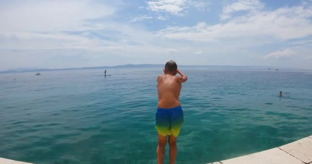 Back view of boy in swim shorts during jumping somersault from pier in clear blue sea, stones can be seen bottom of sea. Summer vacation, lifestyle concept — Stock Video