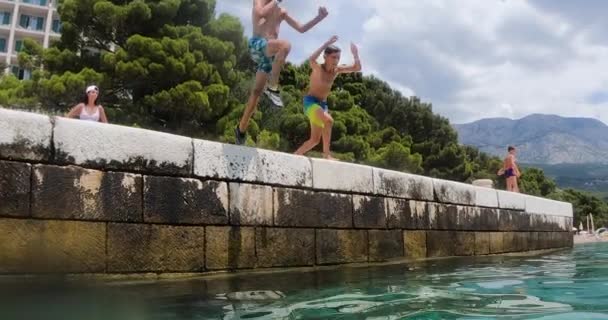 Two slender caucasian boys jumping from pier into clear sea and dive during summer holiday on blue sky and mountains background. — Vídeos de Stock