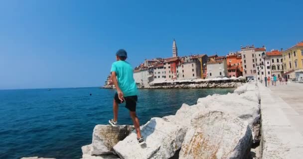 Gyönyörű fiú turista séta a vízparton, kilátással a távolság Adriai-tenger óvárosában Rovinj a nyári vakáció alatt. — Stock videók