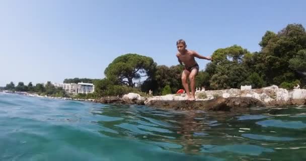 Sports male teenager 10 years in swim shorts jumping from rocky stone into clear Adriatic sea during summer vacation travel. Croatia, Istria — стокове відео
