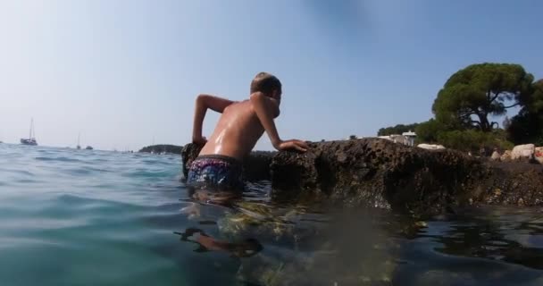 Attraente giovane uomo esce dall'acqua arrampicandosi su pietre marine in mare vicino alla spiaggia al tramonto. Concetto vacanza estiva — Video Stock