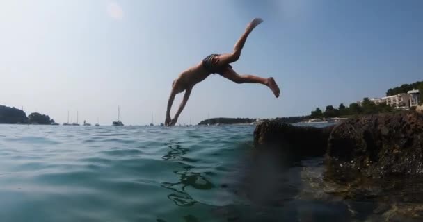 Ung pojke hoppar från klippan, dyka ner i Adriatiska havet vid solnedgången under sommarlovet — Stockvideo