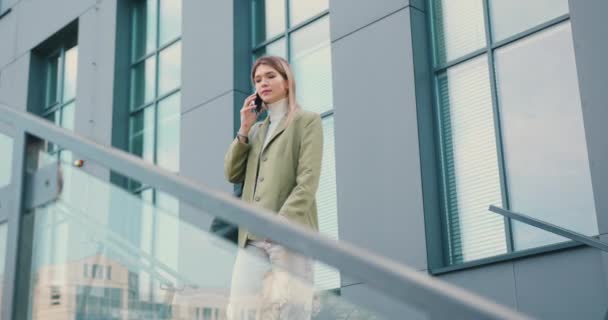 Güzel, genç, gülümseyen bir kadın şehir dışında yürürken akıllı telefondan konuşurken nöbet tutuyor. — Stok video