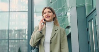 Beyaz kadın cep telefonuyla konuşuyor dışarıda ofisin yanında yürüyor.