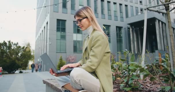 Atrakcyjna młoda kaukaska kobieta pracująca lub ucząca się przy użyciu laptopa siedząc na ławce trawnika kampusu. Pretty girl student przygotowuje się do zajęć na świeżym powietrzu — Wideo stockowe