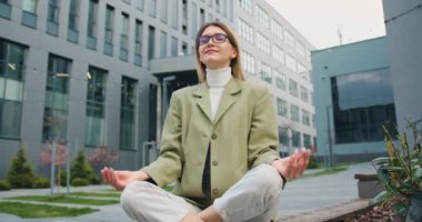 Gözlüklü, gevşemiş genç beyaz bir kadın ofis binasının veya üniversitenin avlusunda lotus pozisyonunda oturuyor. Sağlıklı yaşam tarzı, açık hava.
