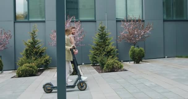 Mujer joven caucásica y hombre caminando por la calle con patada e-scooter y portátil hablando y riendo por los edificios de oficinas modernos. Tecnología, asociación, exterior — Vídeo de stock