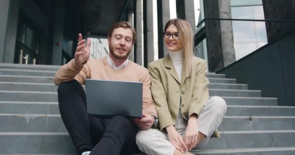 Snygg positiv kaukasisk ung kvinna och man sitter på trappor med laptop, utomhus. Frilansare par sitter i trappor tittar på laptop prata och glädja sig åt framgång — Stockvideo