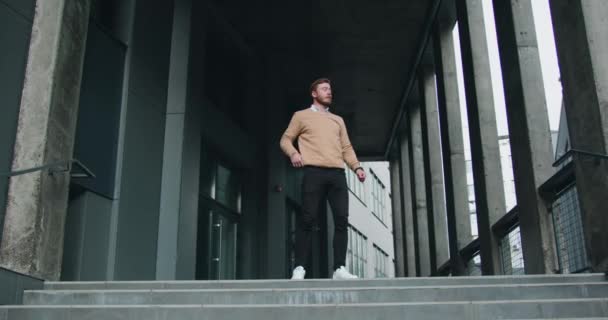 Χαρούμενος και όμορφος καυκάσιος νεαρός γενειοφόρος επιχειρηματίας που στέκεται στα σκαλιά φεύγοντας από το κτίριο του γραφείου και χορεύοντας καθώς βρήκε νέα δουλειά στα όνειρά του. Έξω. — Αρχείο Βίντεο