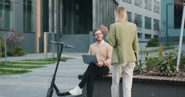 현대 사무실 건물 옆 의자에 앉아 있는 매력적 인 한 쌍의 남녀의 모습, 스마트 한 일상적 인 사업 스타일로 함께 노트북으로 작업하며 웃고 있는 모습. 밖에 는 프리랜서 노동자 개념 — 비디오
