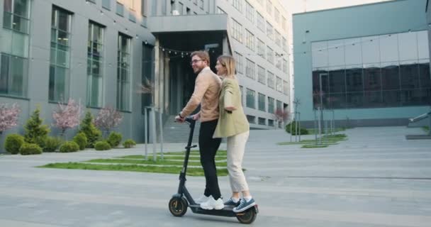 Junges fröhliches kaukasisches Paar, das bei sonnigem Wetter im Stadtpark in der Nähe eines modernen Bürogebäudes Spaß beim Fahren mit dem Elektroroller hat. Lebensstil. Draußen — Stockvideo