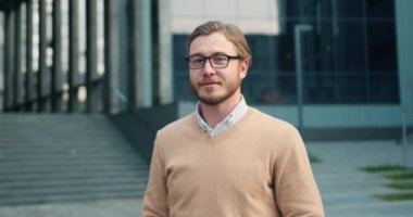 Modern ofis binasının önünde dikilirken kameraya gülümseyen gözlüklü genç iş adamı. Şık giyinmiş pozitif erkek portresi.