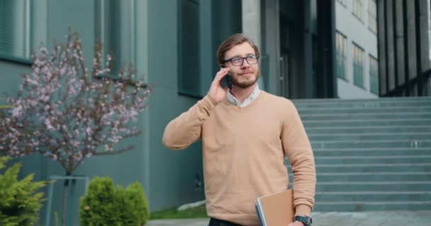 Портрет успешного молодого бизнесмена, одетого в элегантный деловой стиль, выходящего за пределы офисного здания с папкой и разговаривающего по мобильному телефону — стоковое видео
