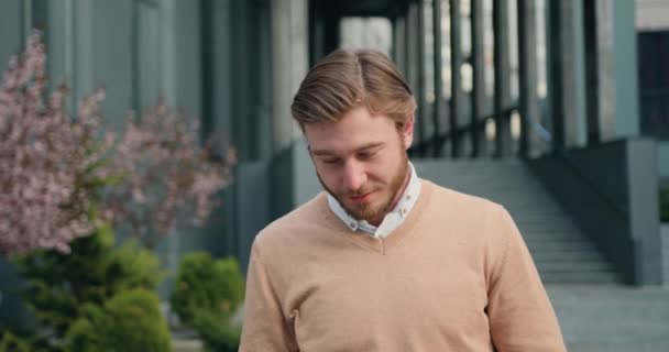 Detailní portrét pohledného úspěšného muže, vzhlížejícího ke kameře, usmívajícího se na pozadí kancelářské budovy. Venku — Stock video