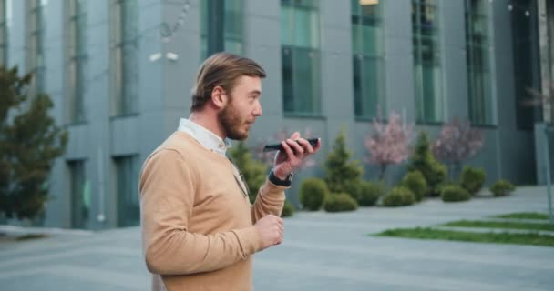 Side view of bearded smiling handsome man talking at mobile phone walking in the street to the office building. Outdoors — Stock Video